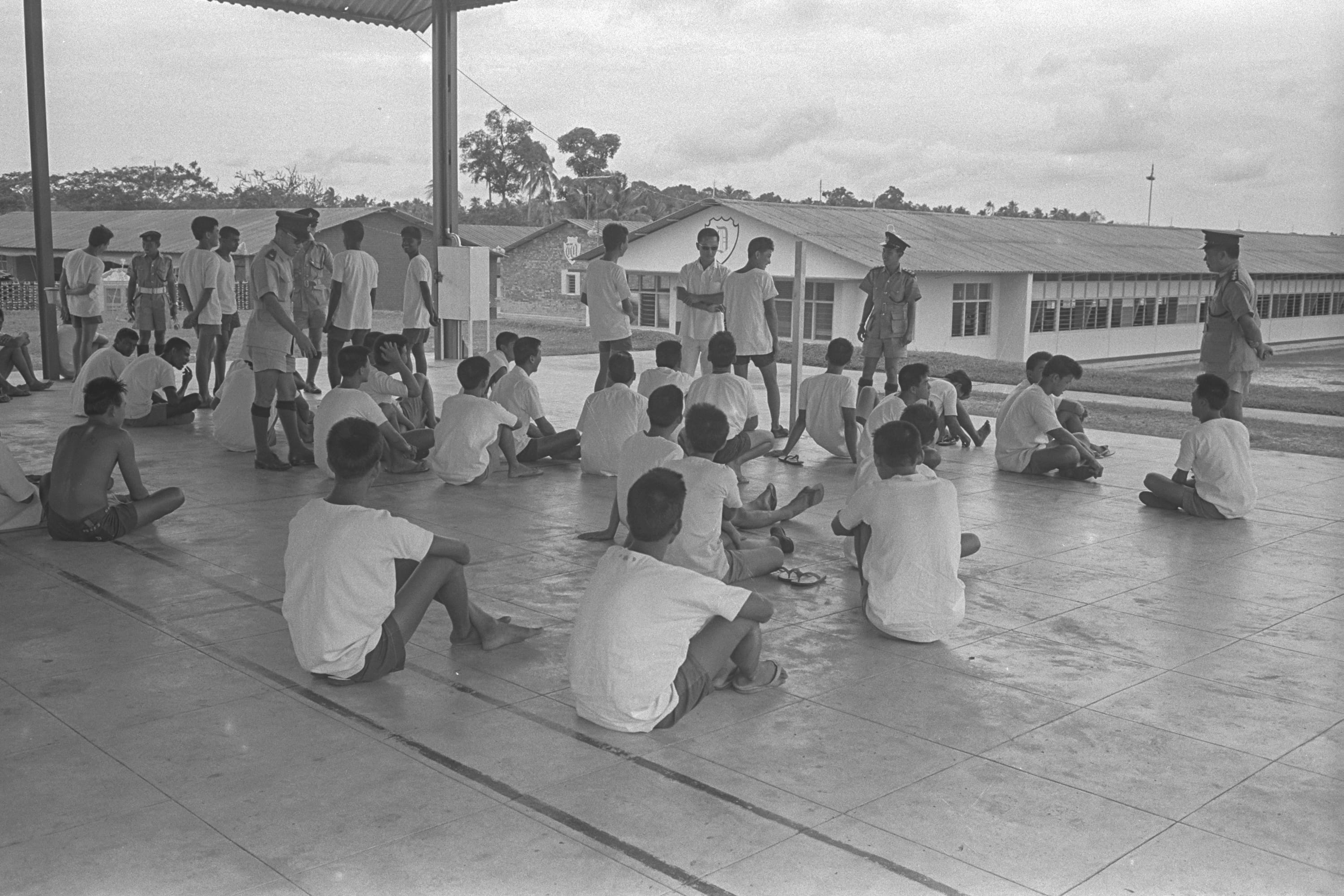 A group of men wearing white shirts and shorts sit on the floor while several uniformed officers stand around them.